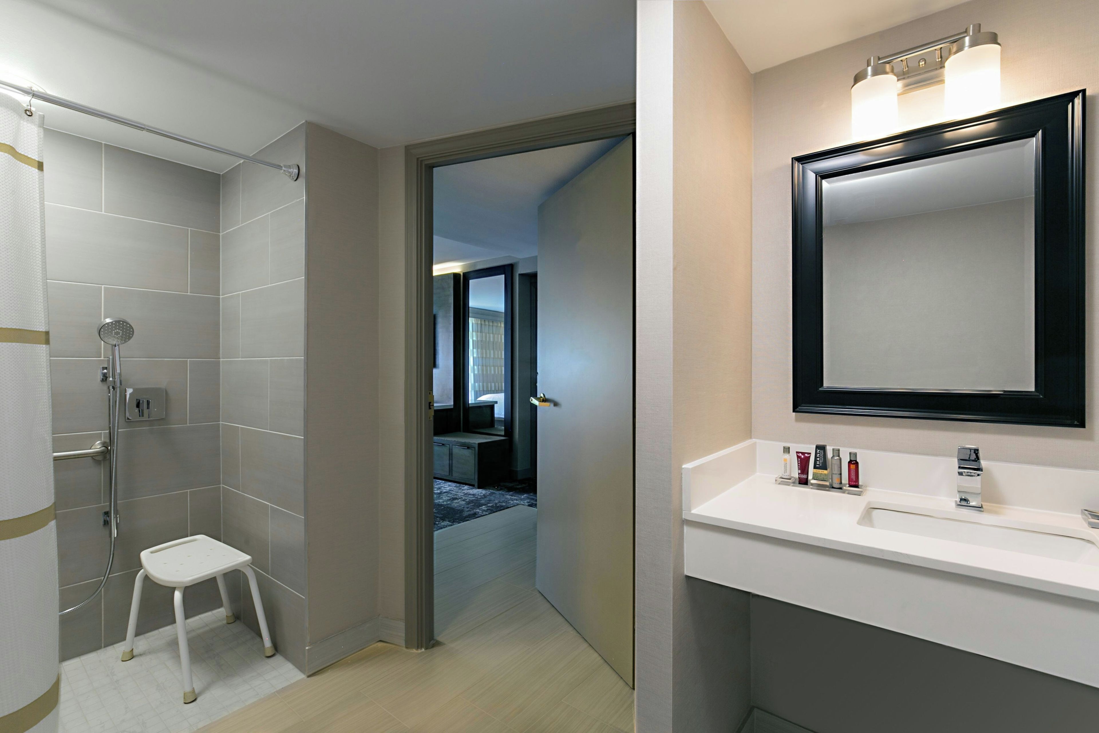 A modern bathroom with a shower, a small stool, and a vanity with a mirror and toiletries.