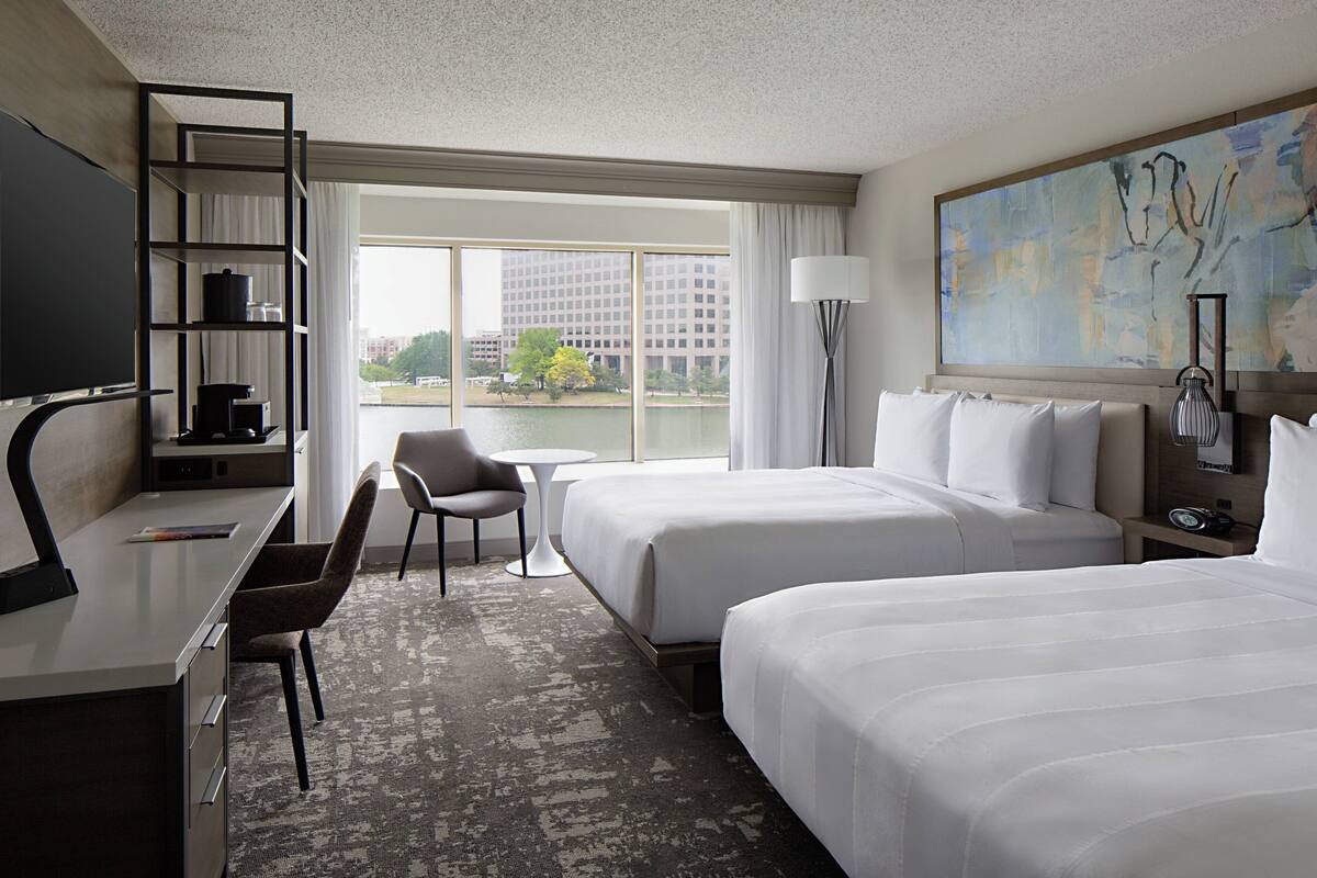 A hotel room with two beds, a desk, a chair, a TV, and a large window with a view of a building and trees.