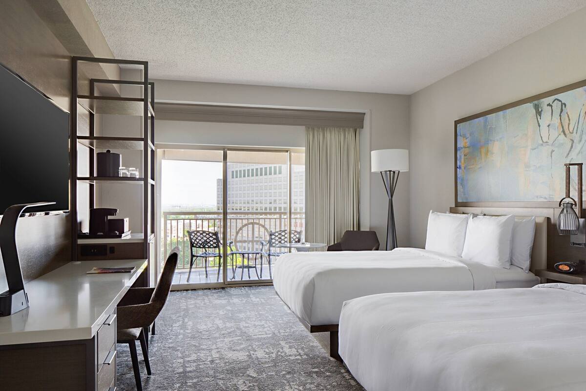 A hotel room with a desk, two beds, a chair, a lamp, and a painting.