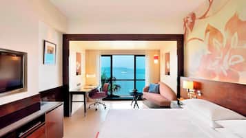 A hotel room with a large bed, a desk, a chair, a TV, and a view of the sea.