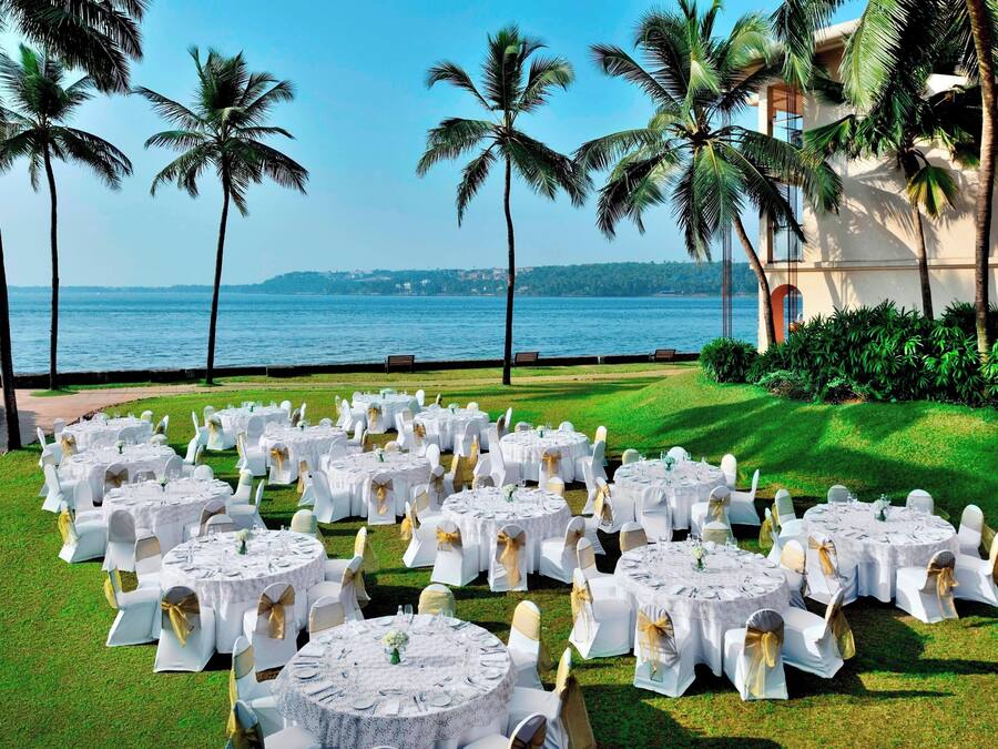 A beachfront venue with round tables set up for an event, surrounded by palm trees and a view of the sea.