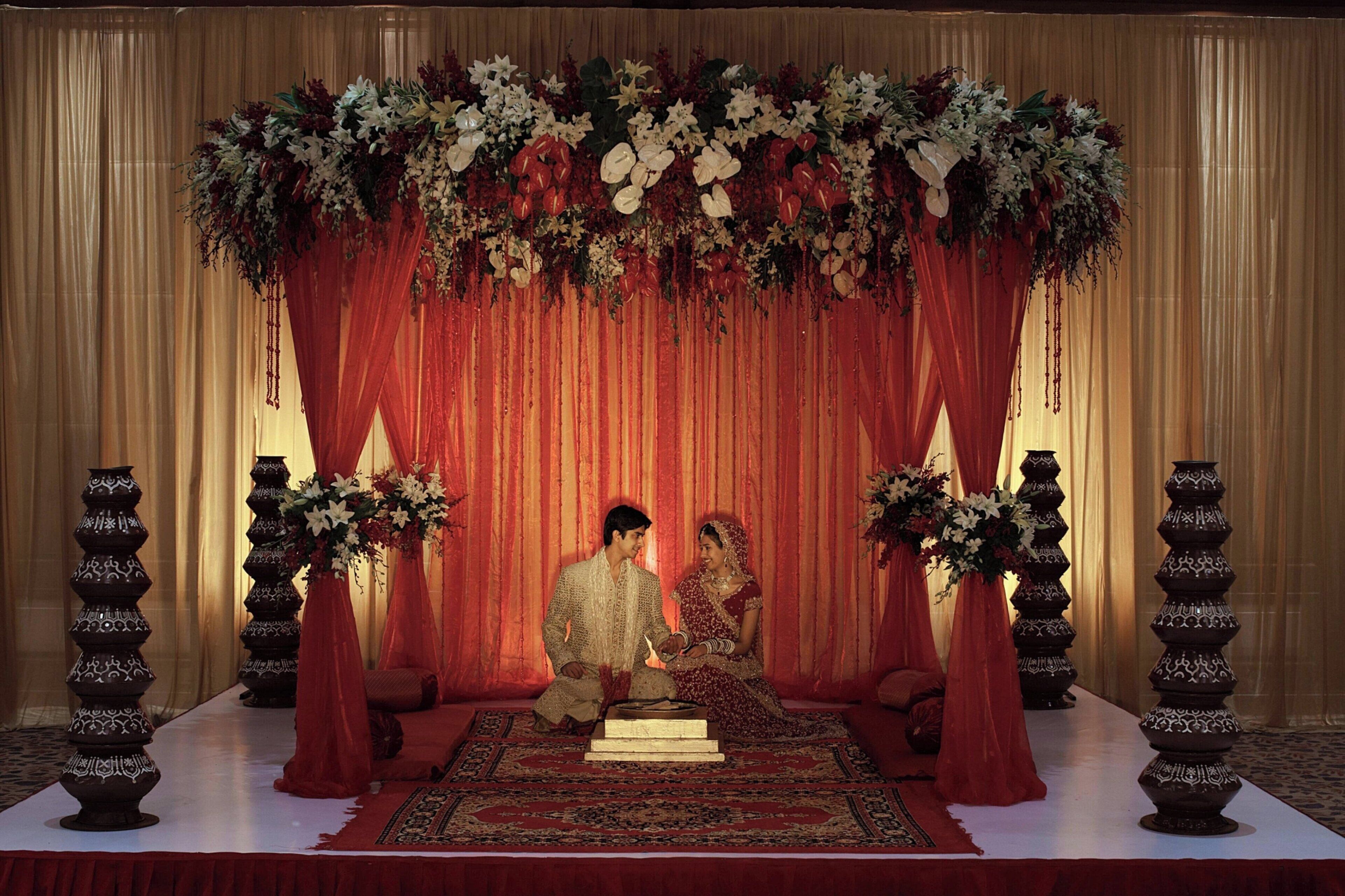 A staged wedding setup with a red draped backdrop, floral arrangements, and traditional vases.