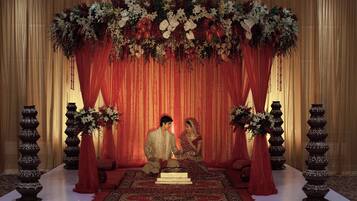 A staged wedding setup with a red draped backdrop, floral arrangements, and traditional vases.