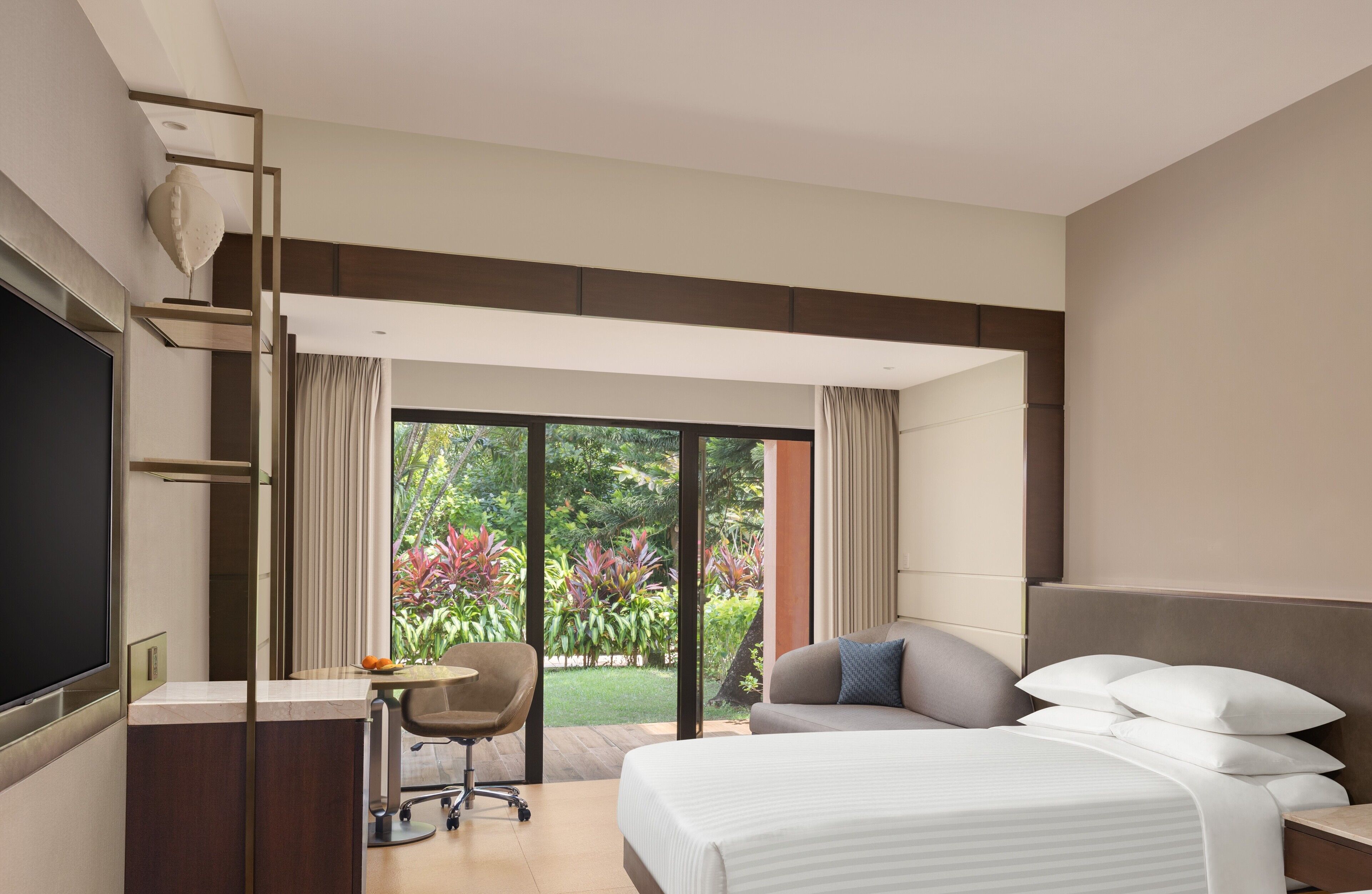 Une chambre d’hôtel avec un lit, un bureau et une chaise. Une grande fenêtre offre une vue sur la verdure environnante.