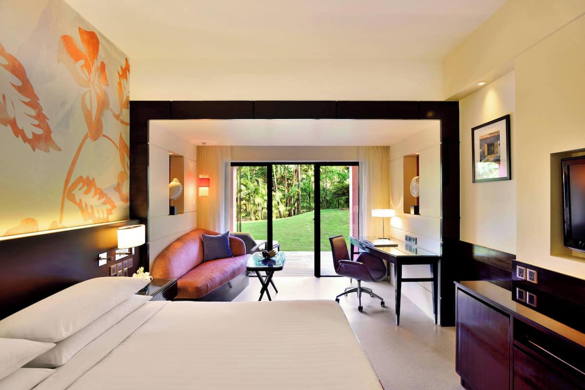 Une chambre d’hôtel avec un grand lit, un bureau, un canapé et une vue sur un jardin verdoyant, visible à travers une porte vitrée.