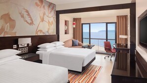 A hotel room with two beds, a desk, a TV, and a large window with a view of the sea.