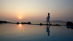 A person walking along a reflective pool at sunset with a view of the ocean and distant hills.