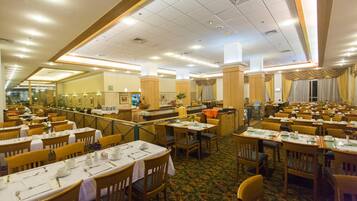 A large dining hall with multiple tables and chairs, a central serving area, and a view of the kitchen in the background.