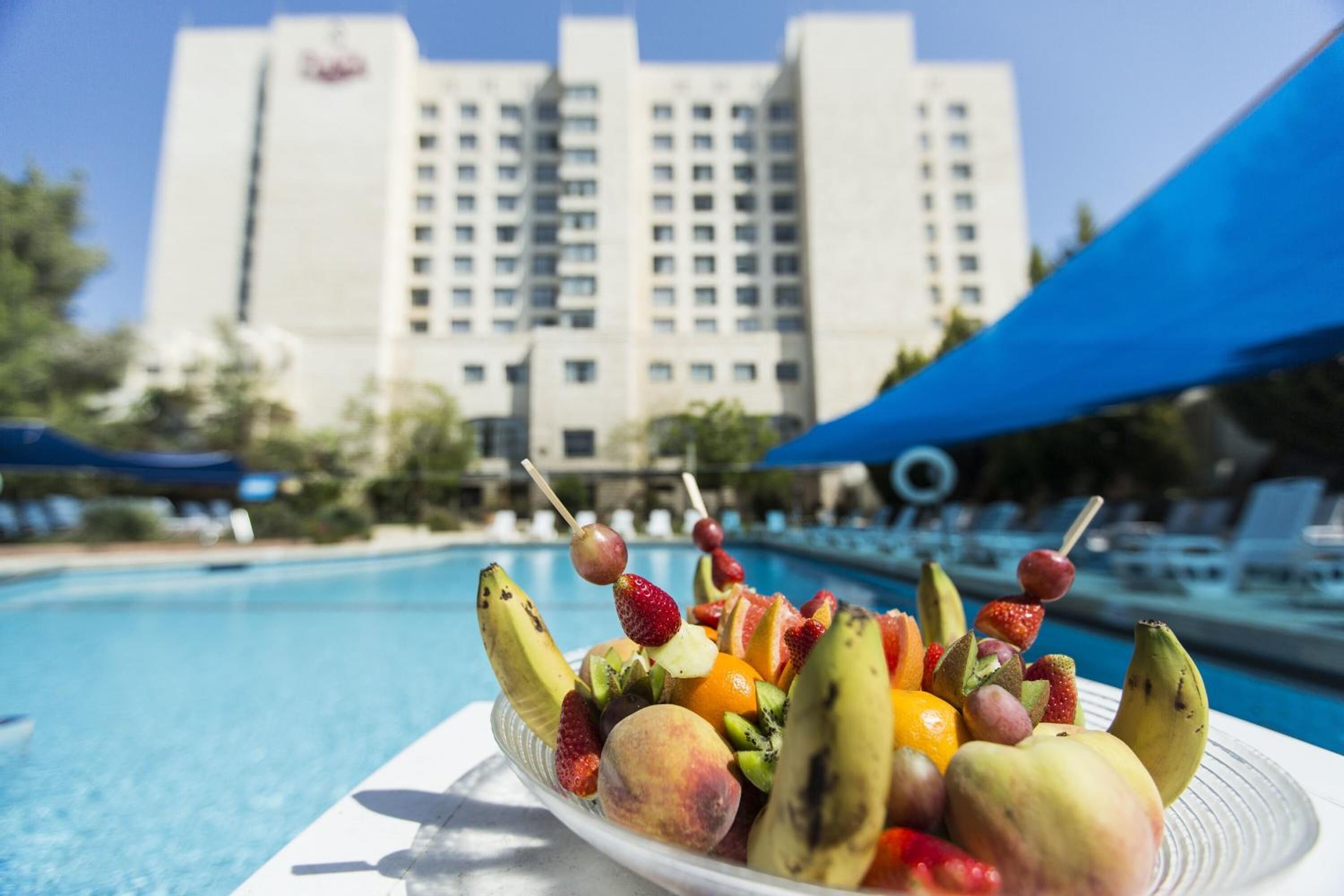 A fruit platter with skewers on a table by a poolside, with a hotel building in the background.