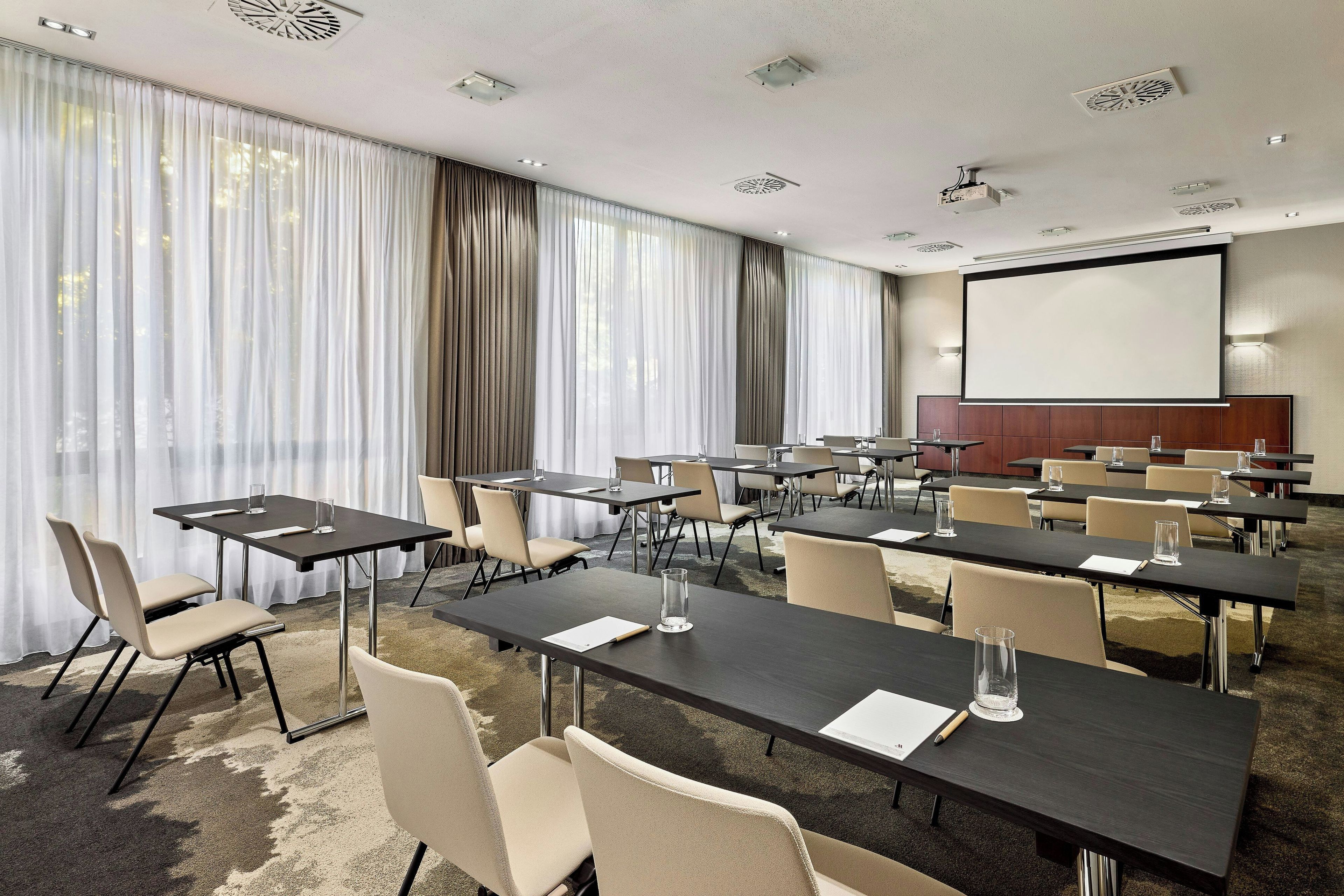 Une salle de conférence équipée de plusieurs tables et chaises, d’un écran de projection et d’une grande fenêtre avec des rideaux.