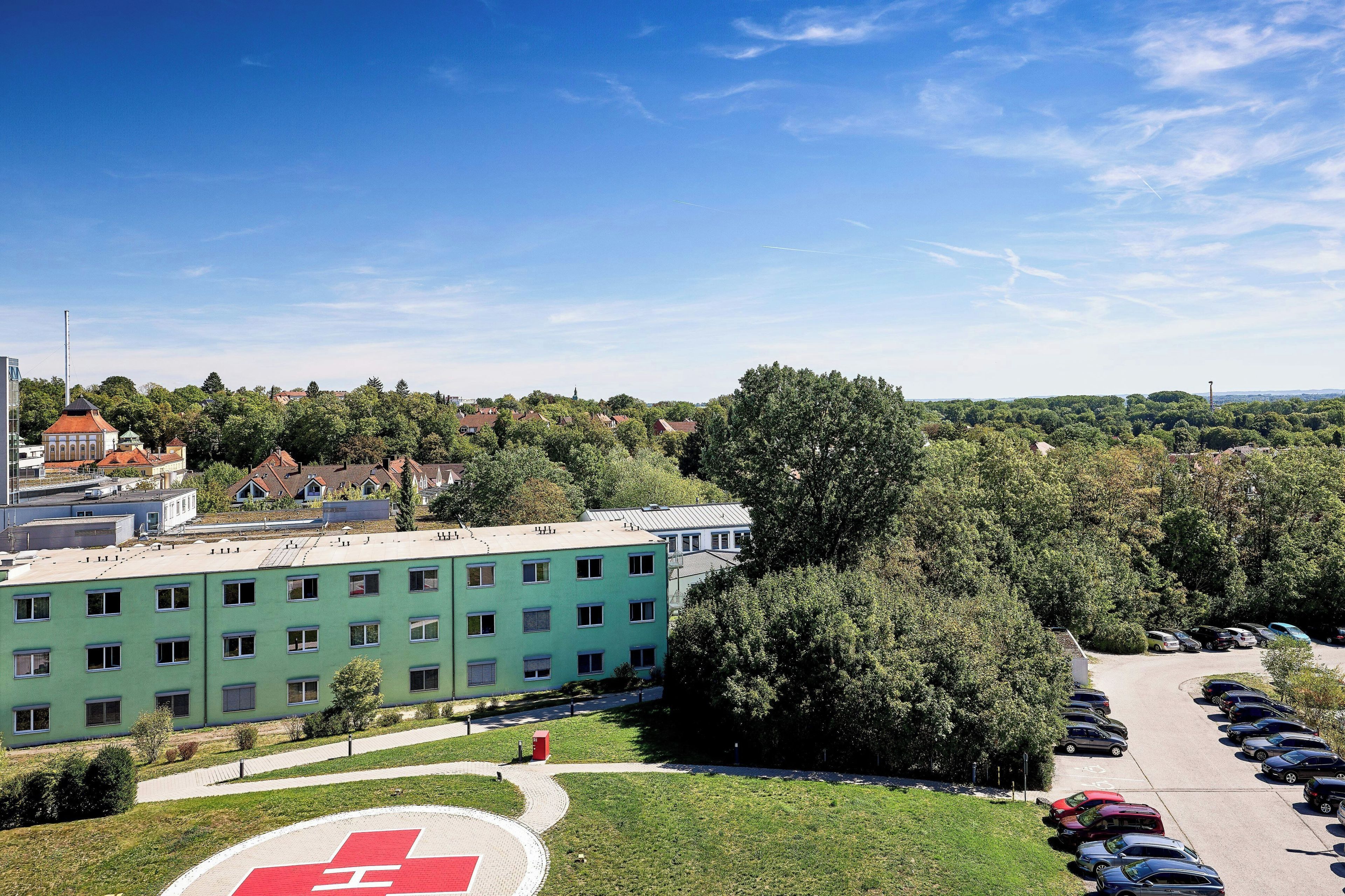 Un bâtiment vert doté de nombreuses fenêtres, un parking avec plusieurs voitures et un grand arbre au premier plan.