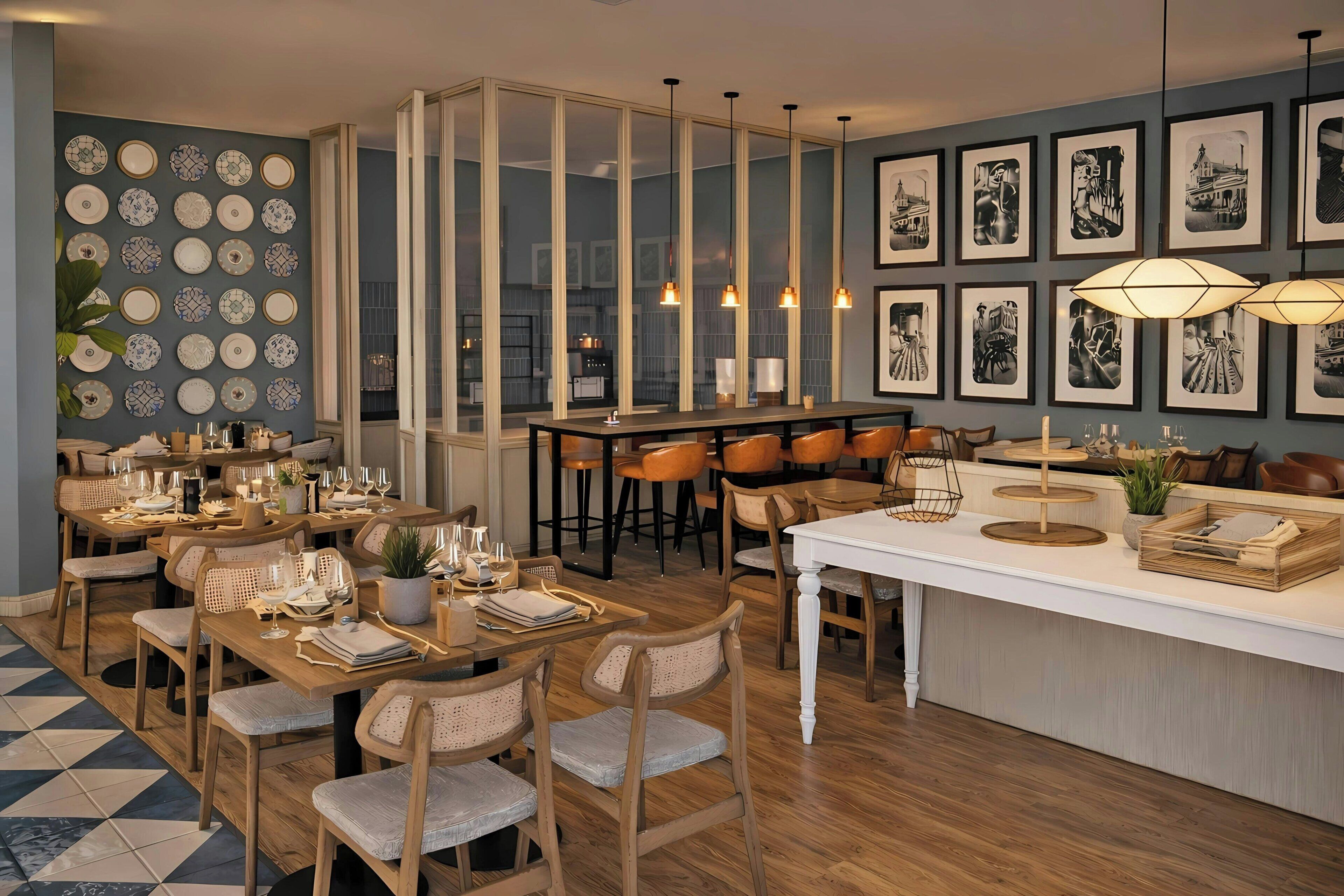 A modern dining area with wooden tables and chairs, a kitchen in the background, and a wall decorated with framed pictures.
