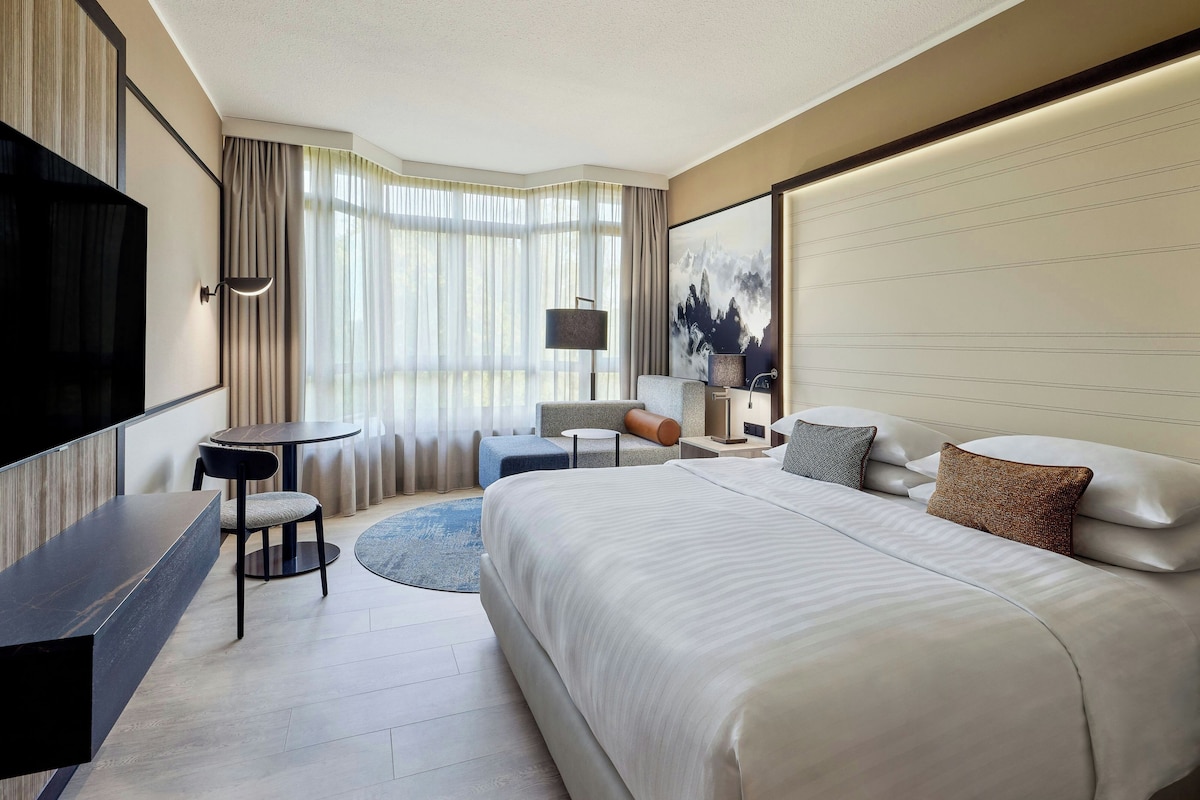 A hotel room with a large bed, a desk, a chair, a small round table, a sofa, and a TV mounted on the wall.