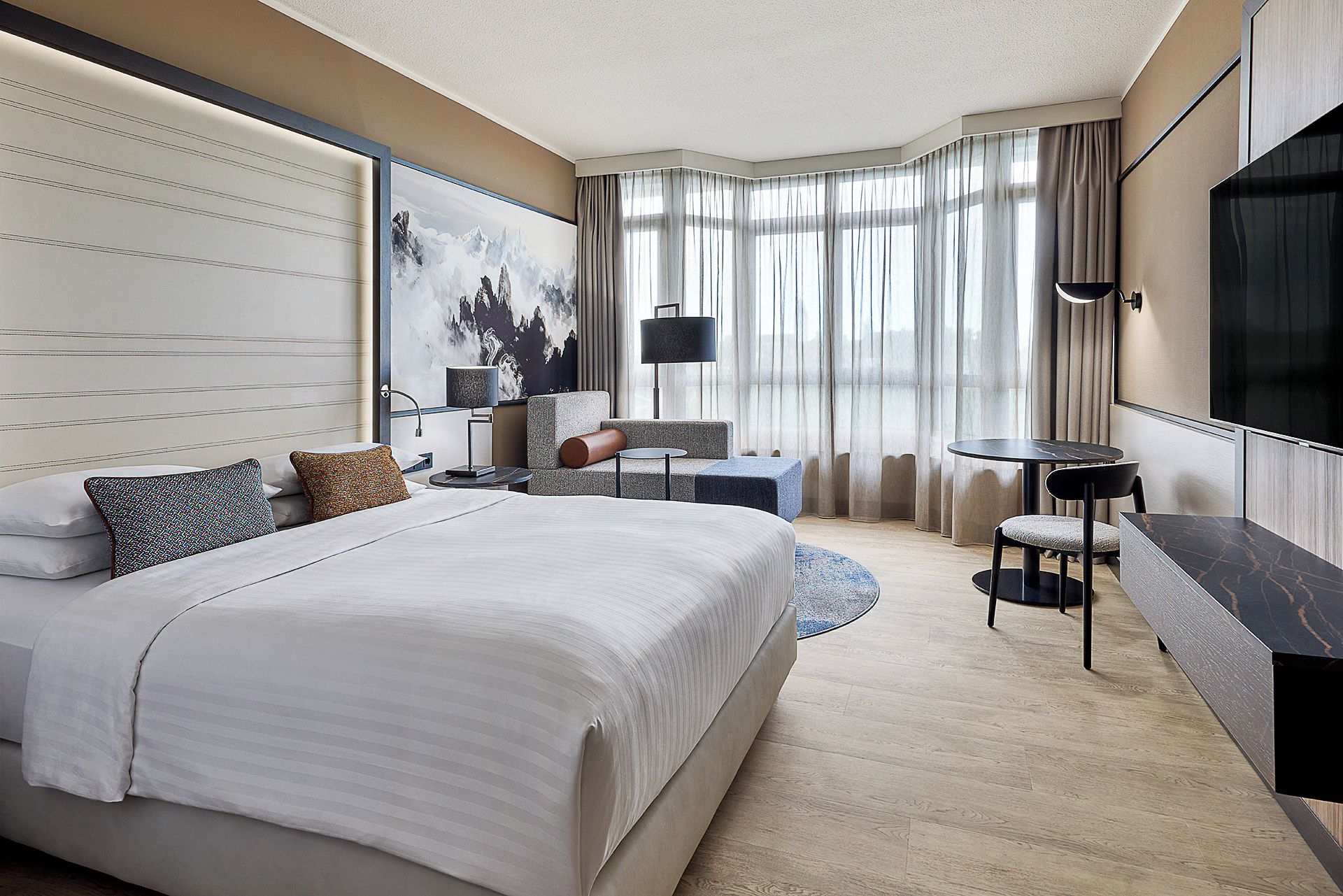 Ein modernes Hotelzimmer mit einem großen Bett, einer Sitzecke mit Tisch, einem Fernseher und einem großen Fenster mit Vorhängen.