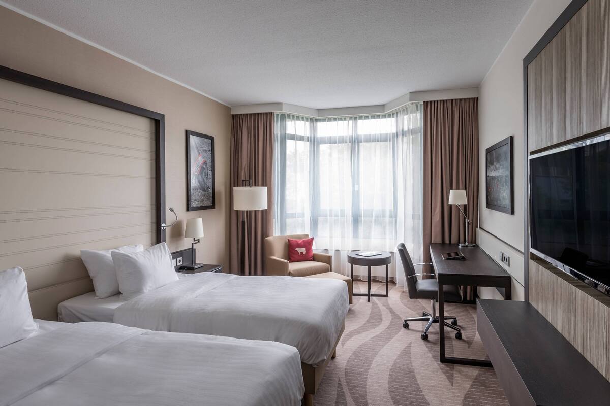 A hotel room with two beds, a desk, a chair, a TV, and a window with curtains.