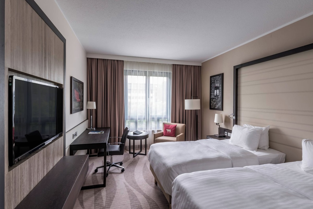 A hotel room with two beds, a desk, a chair, a TV, and a window with curtains.