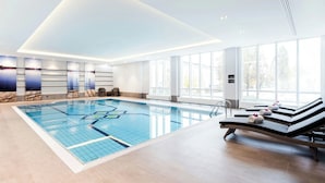 Une piscine intérieure moderne avec une eau d’un bleu limpide, de grandes fenêtres baignées de lumière et des chaises longues avec des serviettes à disposition.