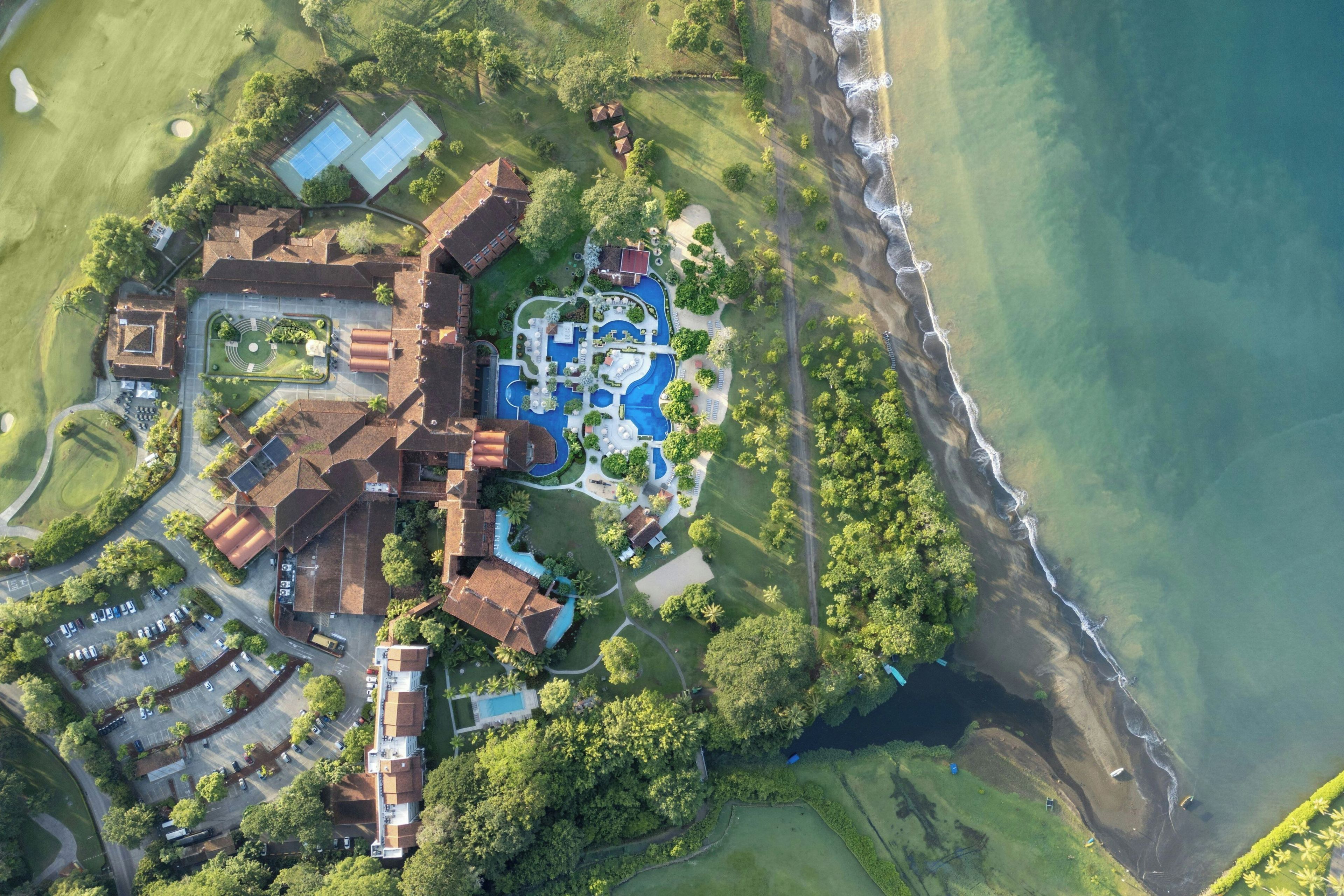 An aerial view of a coastal resort with multiple buildings, a swimming pool area, and a beachfront.