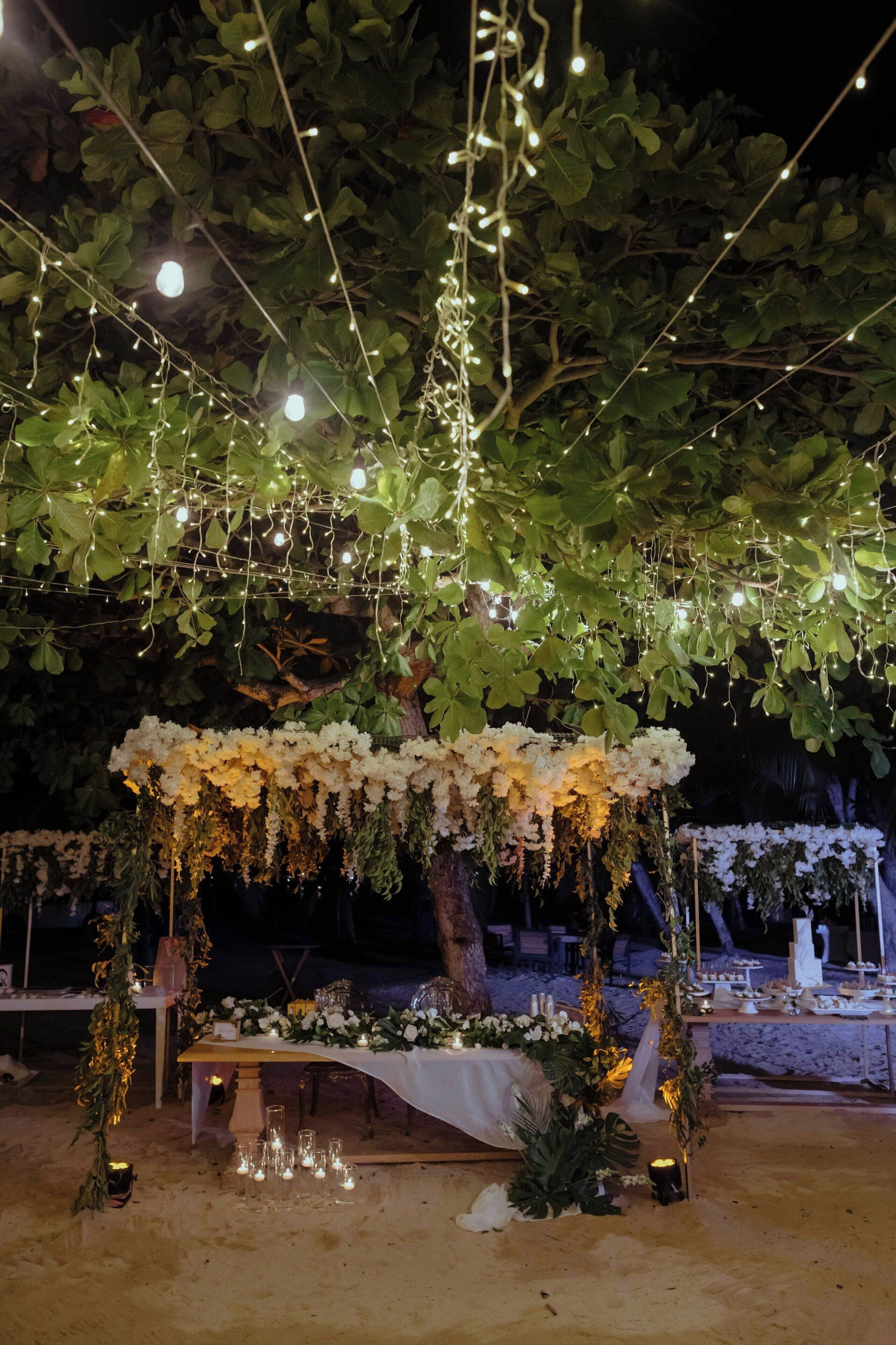An outdoor wedding setup with a canopy of white flowers, string lights, and a table with candles.