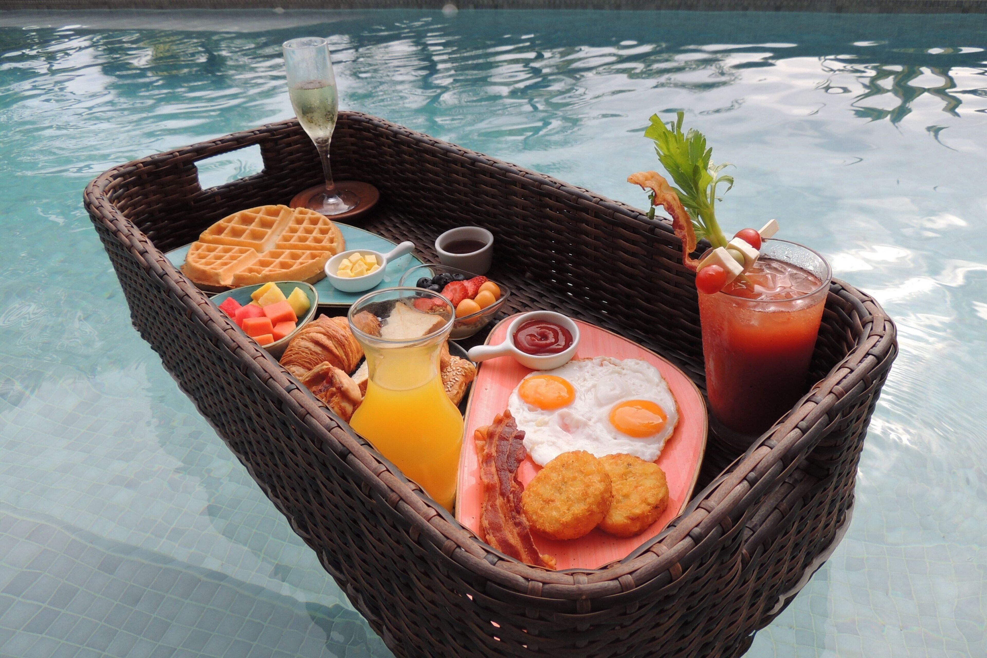 A wicker basket filled with breakfast items including waffles, eggs, bacon, toast, and beverages, placed on a poolside table.