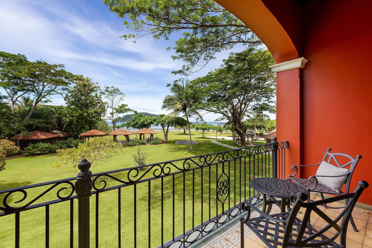 A balcony with a table and chair, a red wall, and a view of a well-maintained lawn and trees.