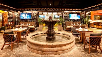 Un restaurante con una fuente central, varias mesas, sillas y televisores en las paredes.