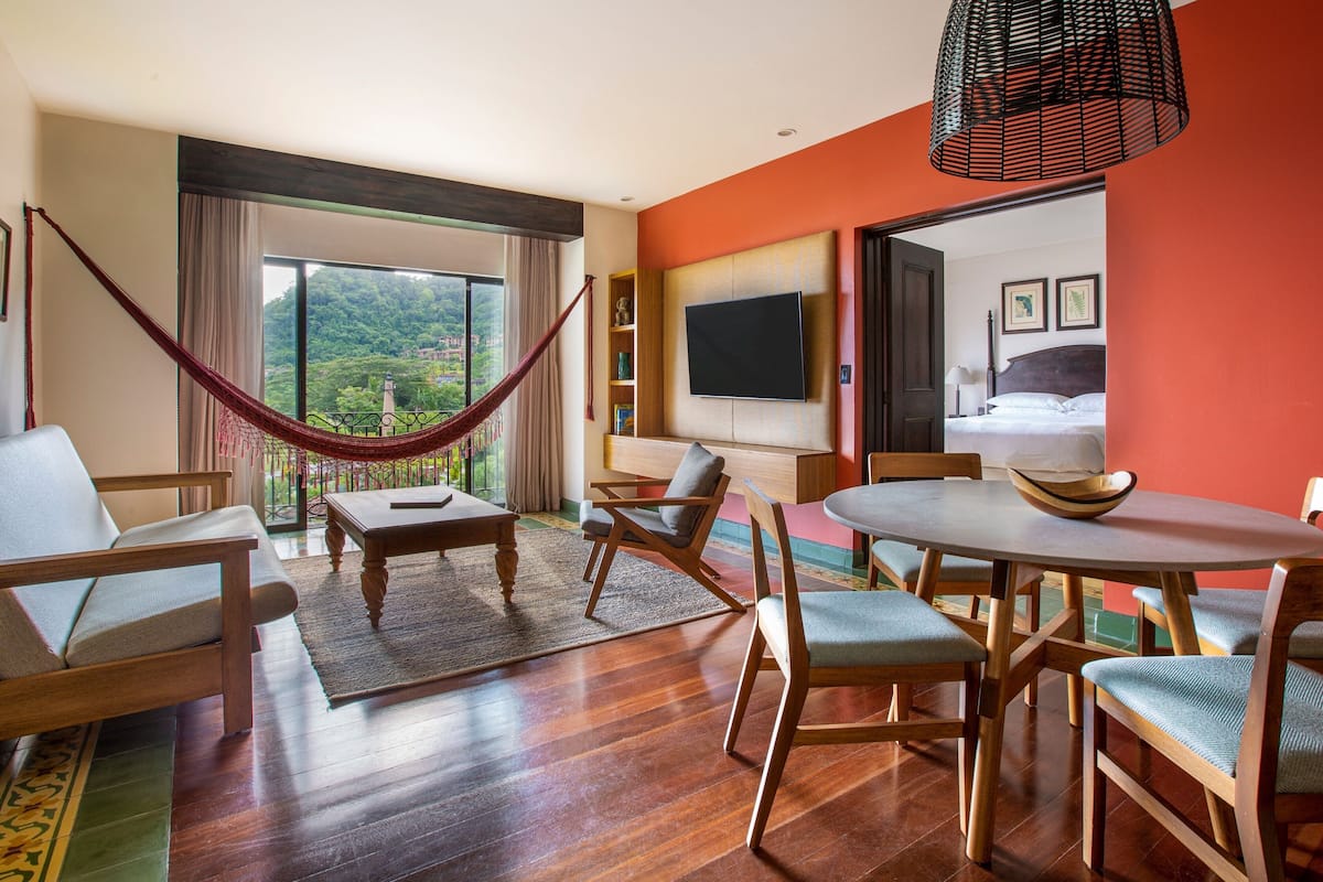 A well-lit room with a large window, a hammock, a dining table with chairs, and a television mounted on the wall.