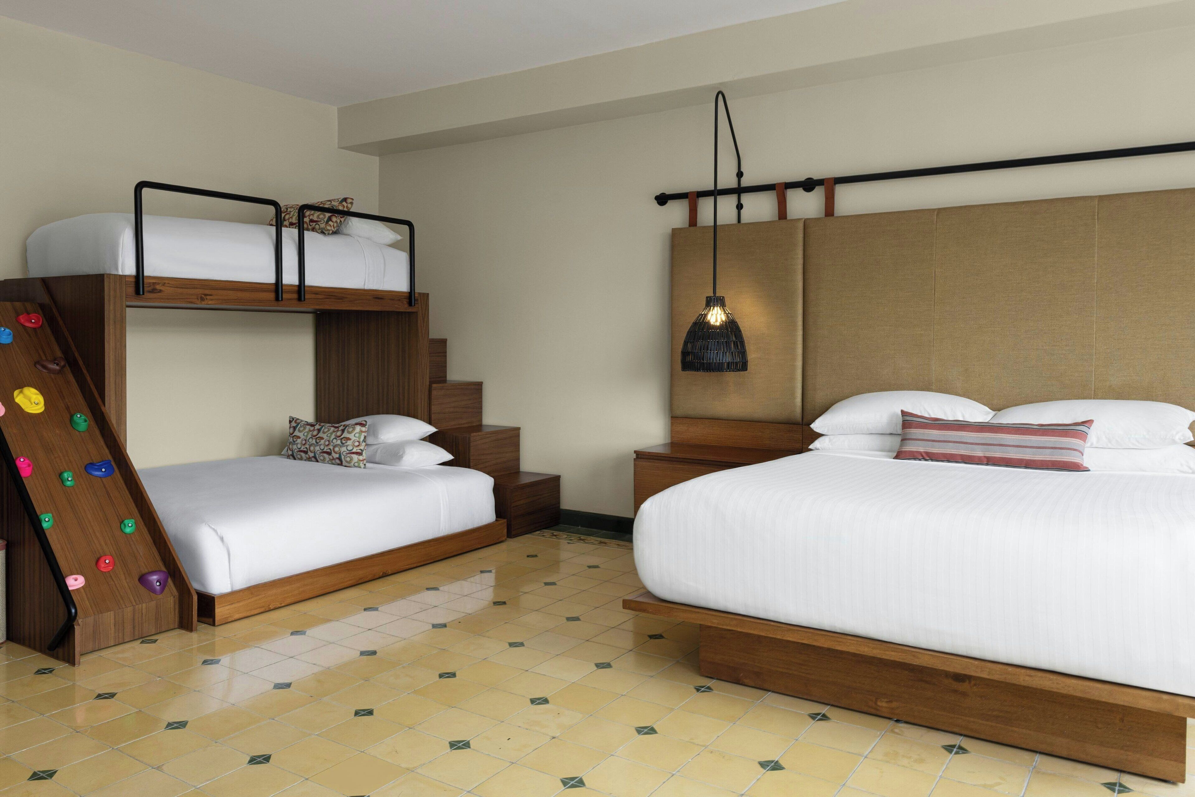 Quarto de hotel com duas camas, incluindo uma cama beliche de madeira, uma parede de escalada e um lustre.