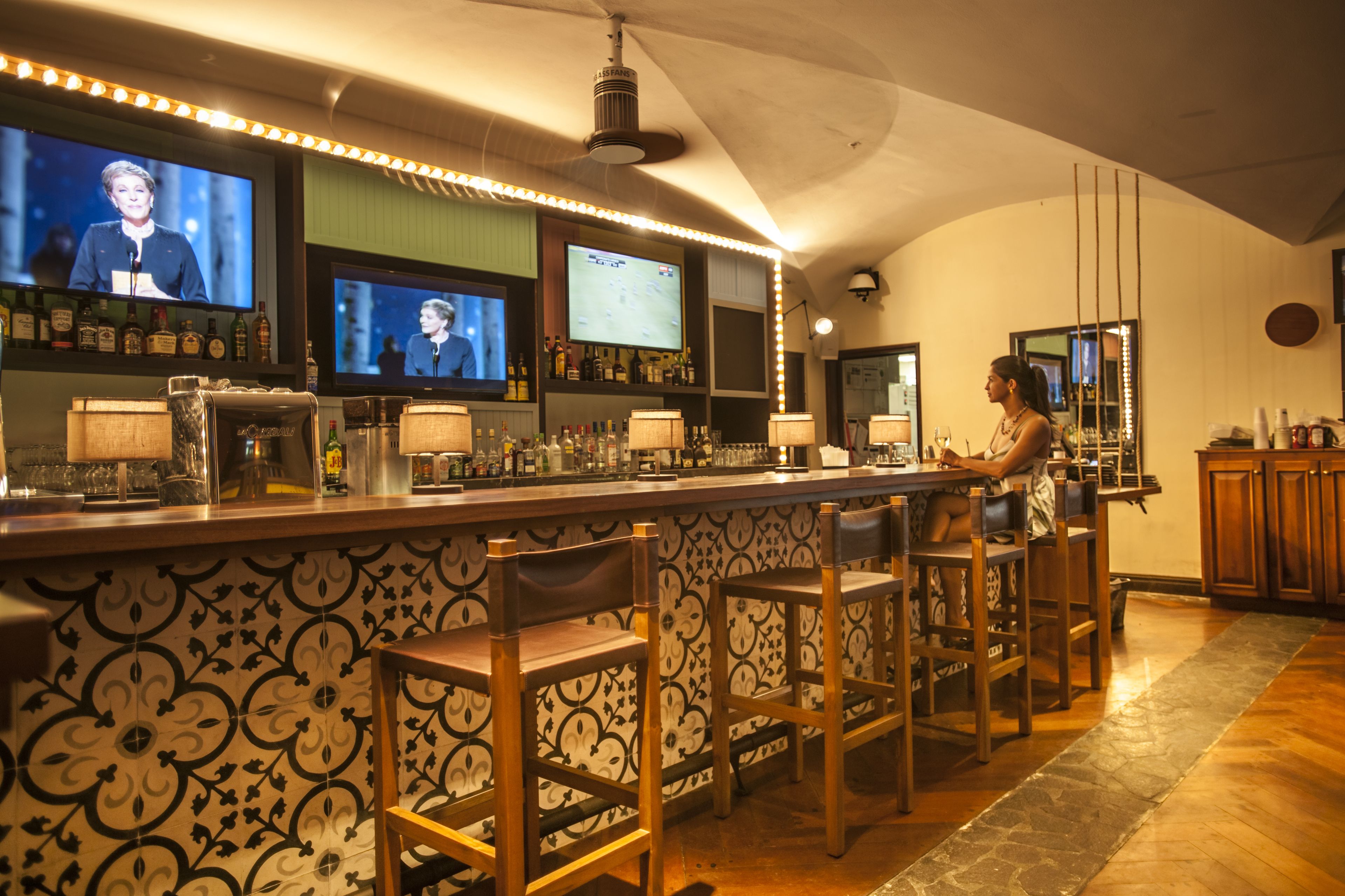 Un espace bar avec un mur décoré de motifs, des tabourets de bar et des téléviseurs diffusant les actualités.