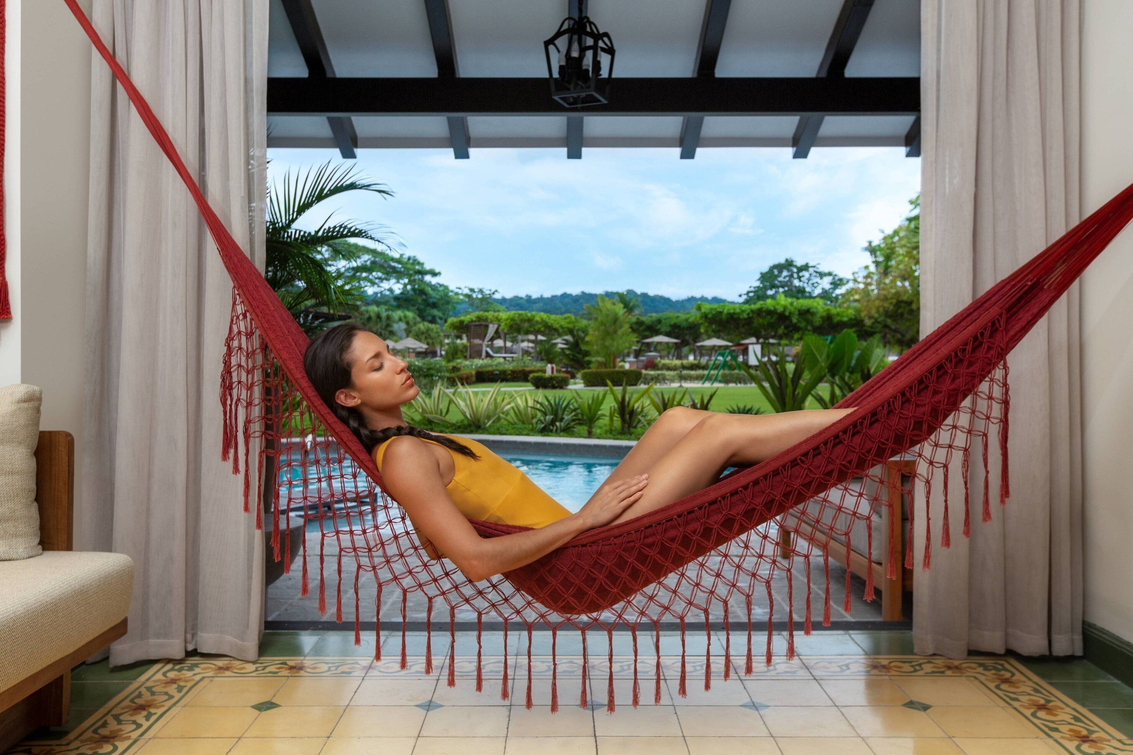 A person relaxing in a red hammock by a pool with a view of greenery and houses in the background.