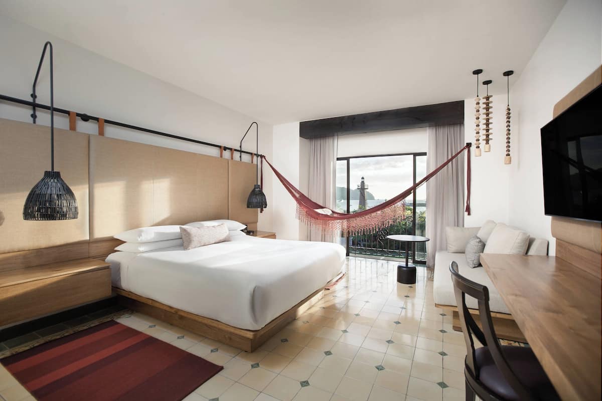 A modern hotel room with a large bed, a desk, a chair, a hammock, and a balcony with a view.
