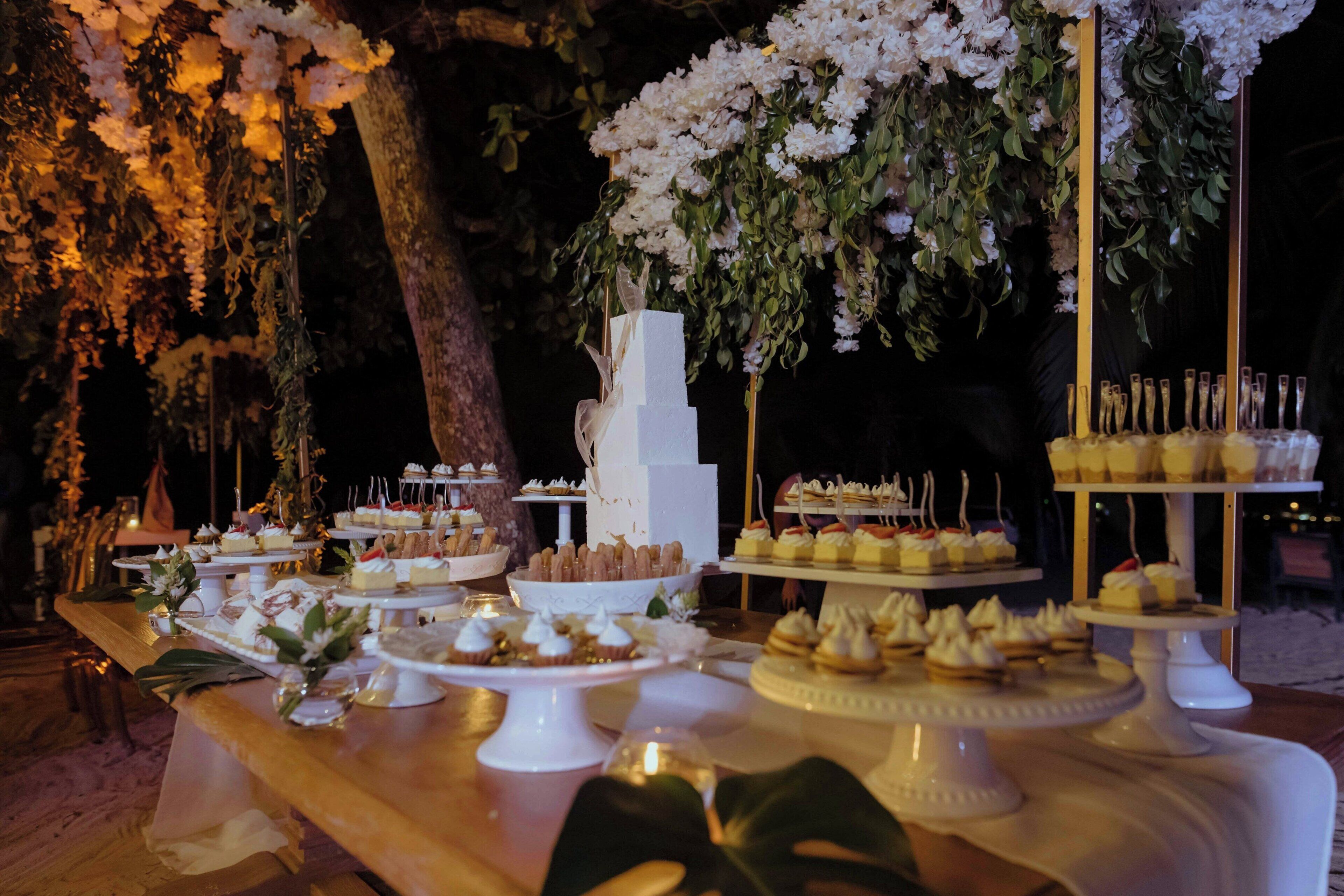 Un tavolo dei dolci con varie paste e torte, con uno sfondo di fiori bianchi e verdeggianti.