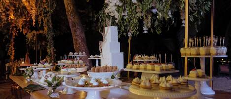 Un tavolo dei dolci con varie paste e torte, con uno sfondo di fiori bianchi e verdeggianti.