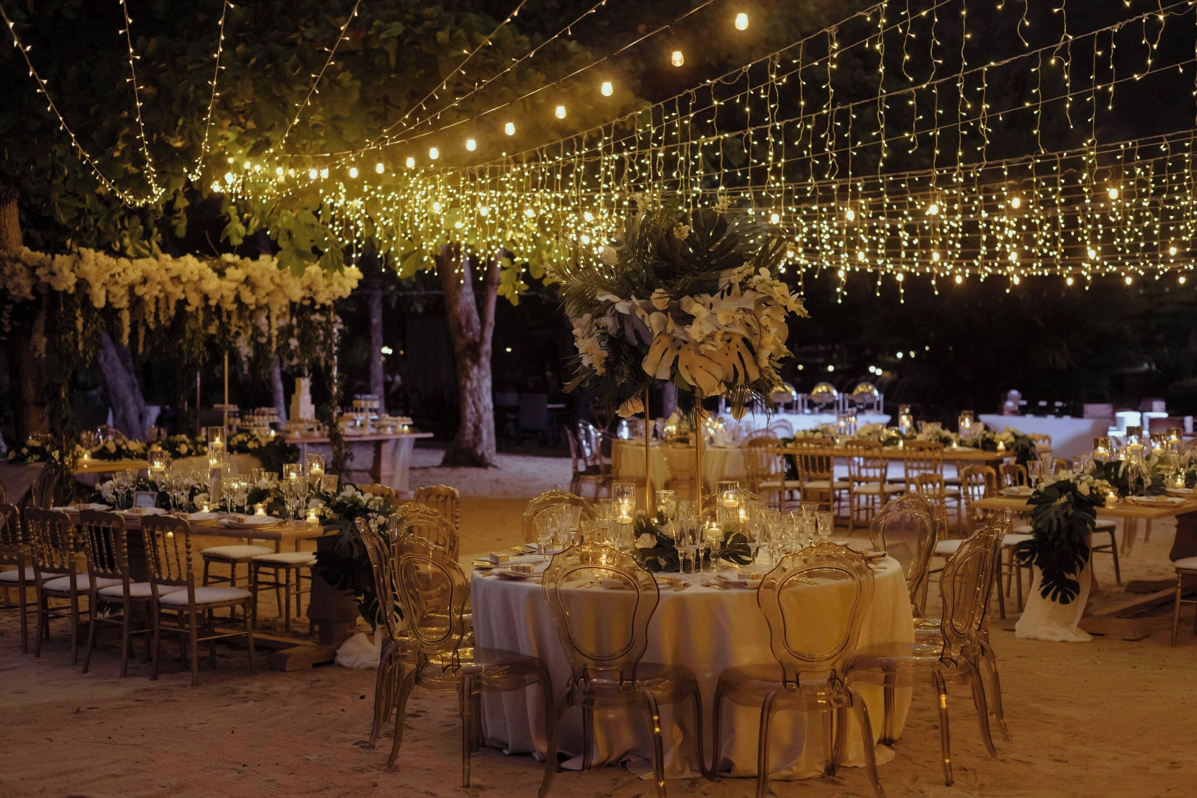 Una recepción de boda al aire libre con mesas redondas, sillas e iluminación de guirnaldas.