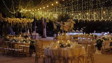 Una recepción de boda al aire libre con mesas redondas, sillas e iluminación de guirnaldas.