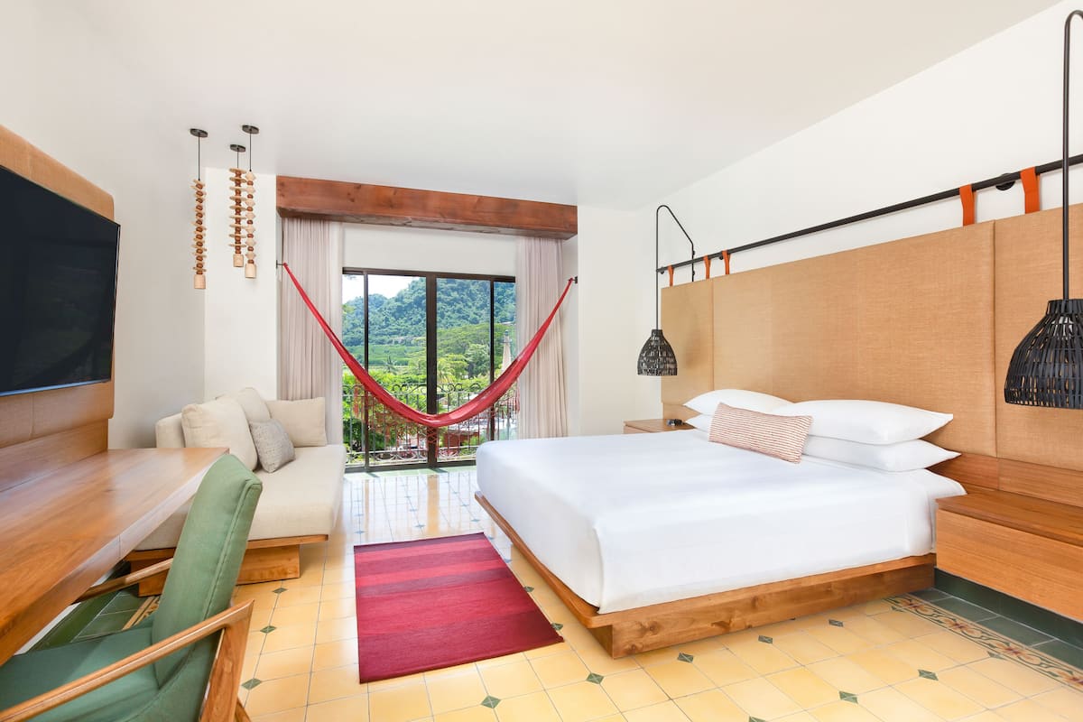 A hotel room with a bed, a desk, a chair, a TV, a hammock, and a view of greenery.