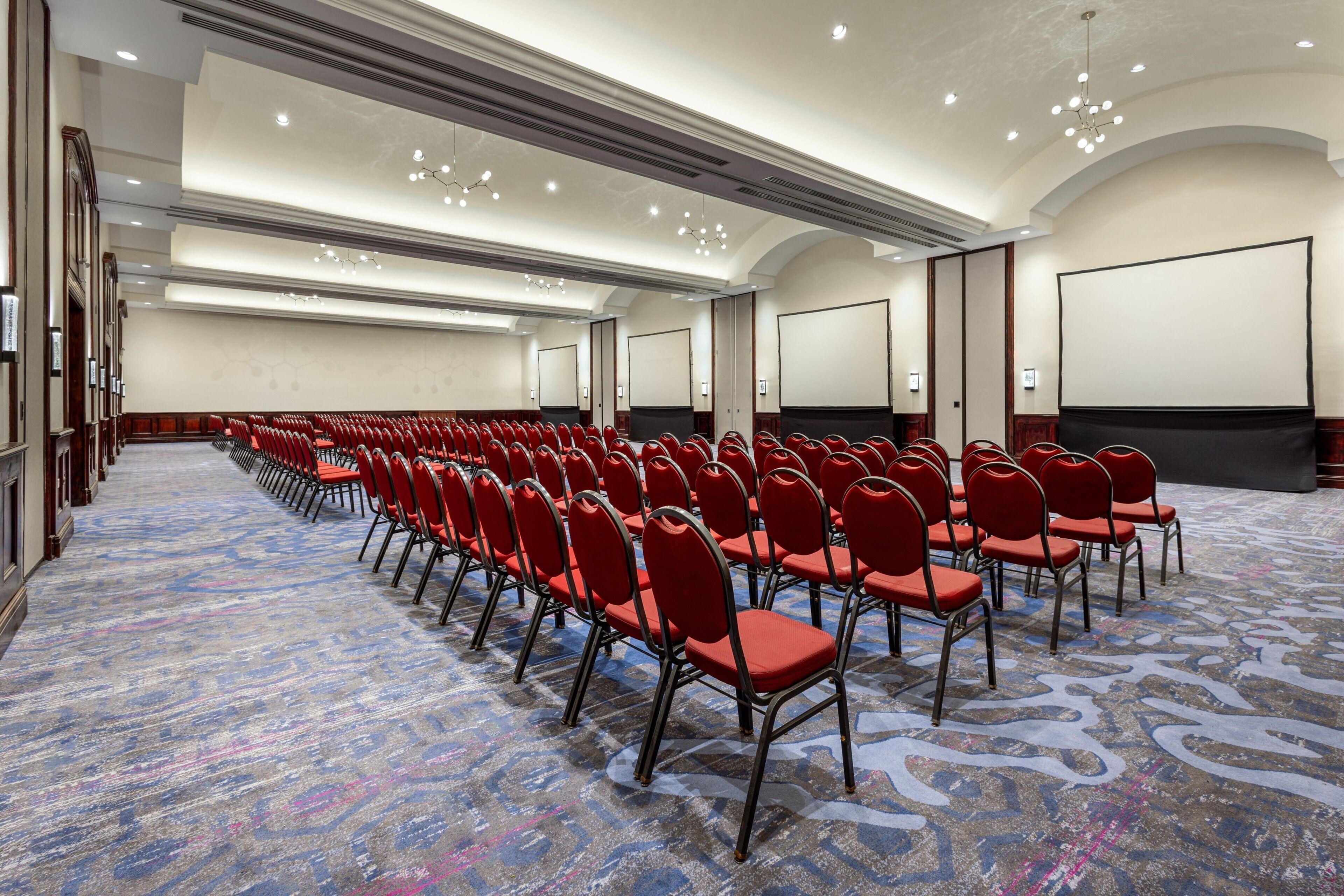 A conference room with rows of red chairs, a patterned carpet, and a large screen at the front.