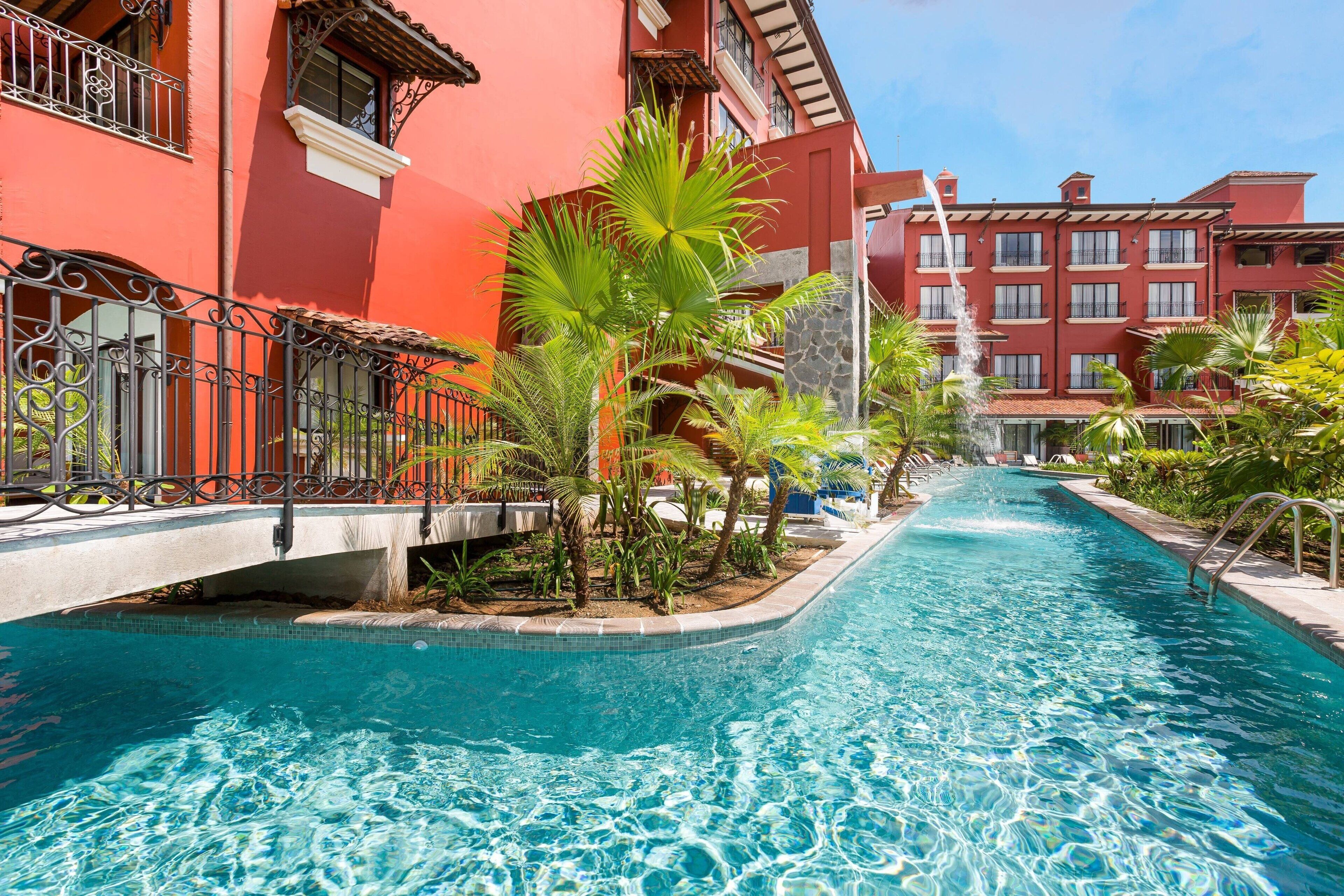 A swimming pool with a clear blue water, surrounded by palm trees and a red building with balconies.