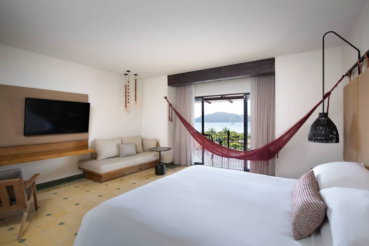 A hotel room with a large bed, a sofa, a TV mounted on the wall, a hammock, and a balcony with a view.