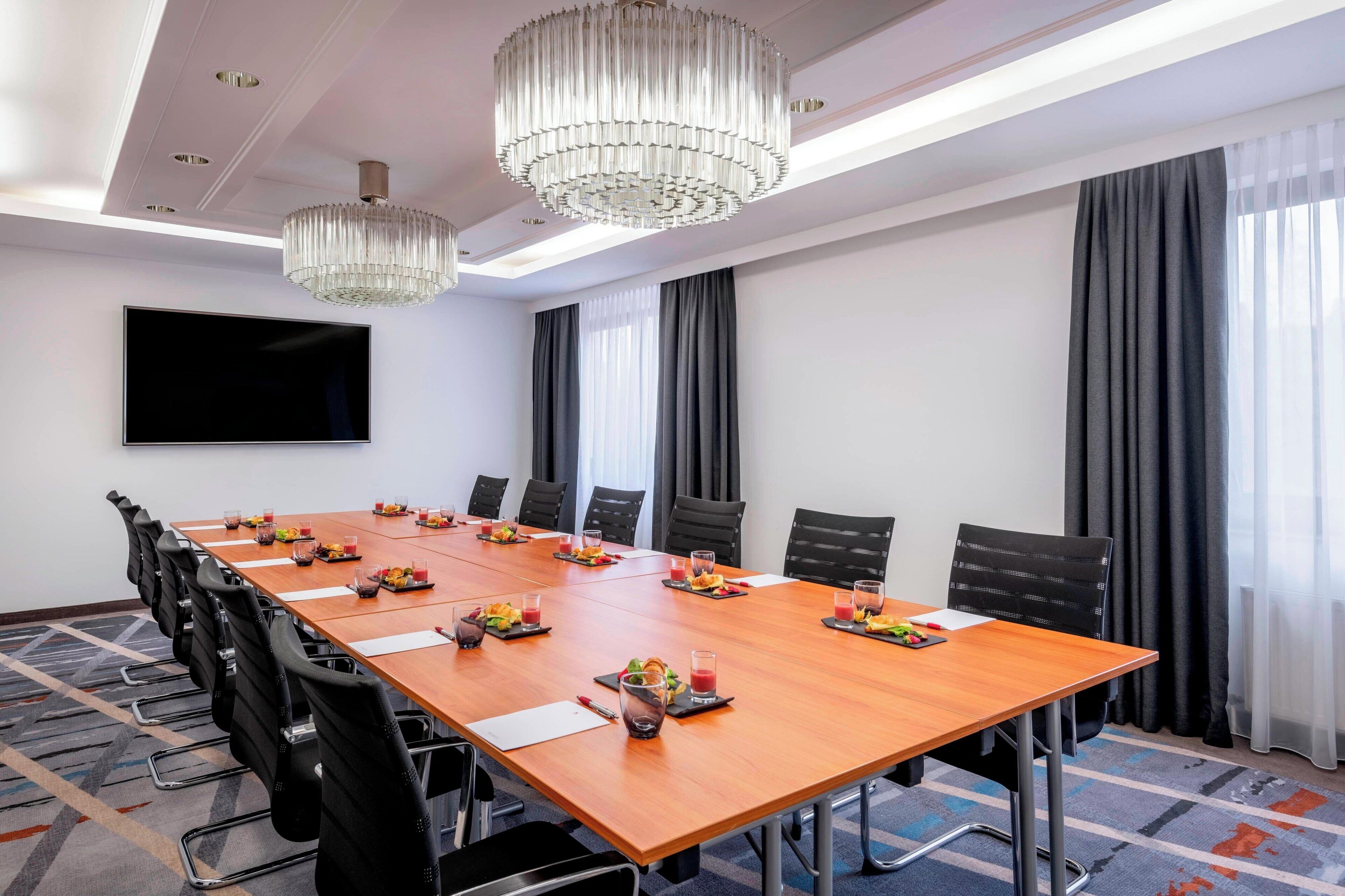 A conference room with a long table, black chairs, a chandelier, a flat-screen TV, and a window with curtains.