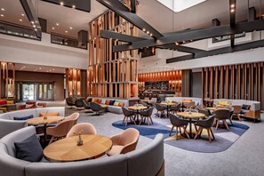 A spacious lobby with multiple seating areas, including sofas and round tables, and a bar area with a high ceiling and exposed beams.