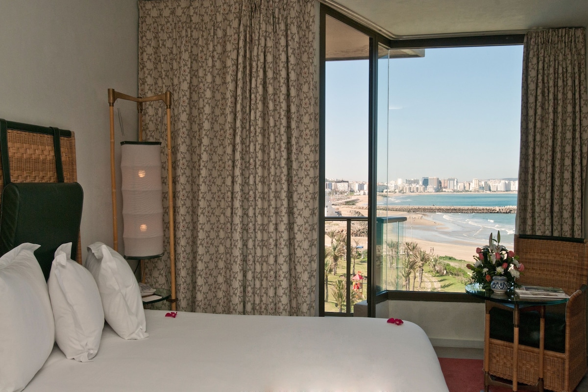 A hotel room with a large bed, a view of the beach, and a balcony with a wicker chair and table.