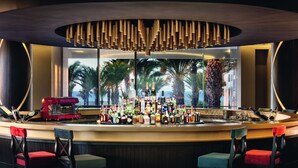 A bar area with a variety of bottles and a coffee machine, surrounded by chairs and a large window with a view of palm trees. - Mövenpick Hotel & Casino Malabata Tanger (Tangier)
