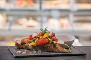 Un sándwich con diversos vegetales sobre una tabla de pizarra, con un fondo desenfocado de otros alimentos.