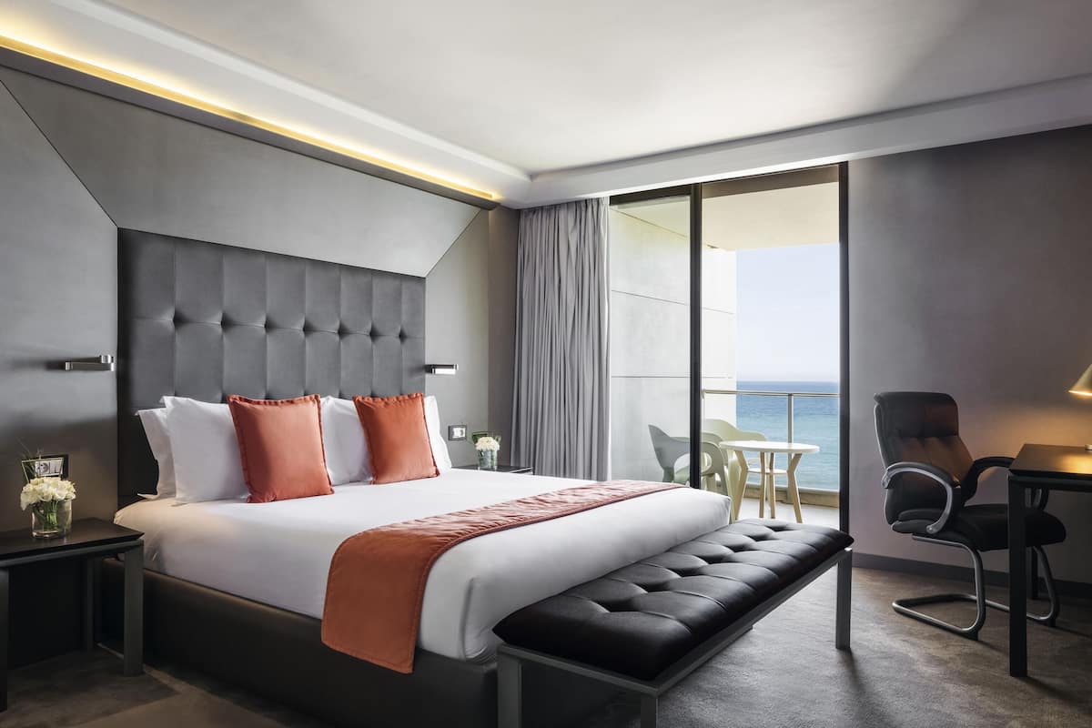 A modern hotel room with a large bed, a bench, a desk, and a view of the sea.