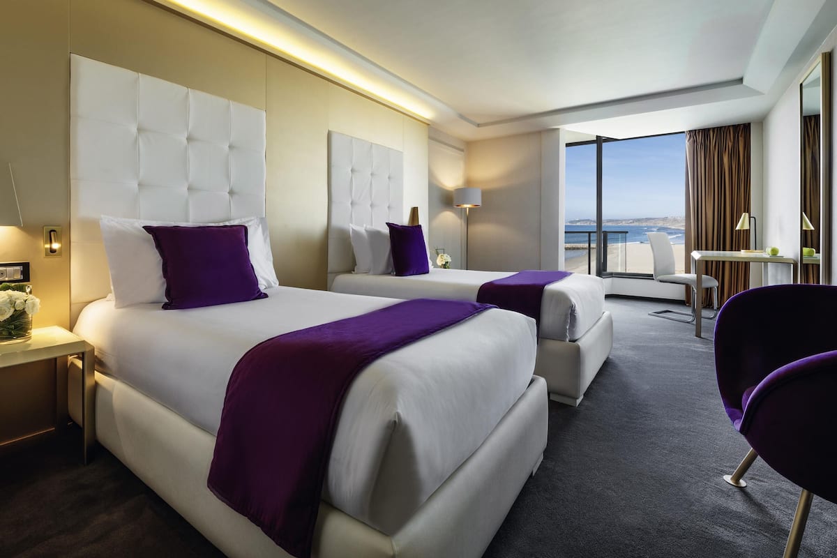 A modern hotel room with two beds, a sitting area, and a view of the sea.
