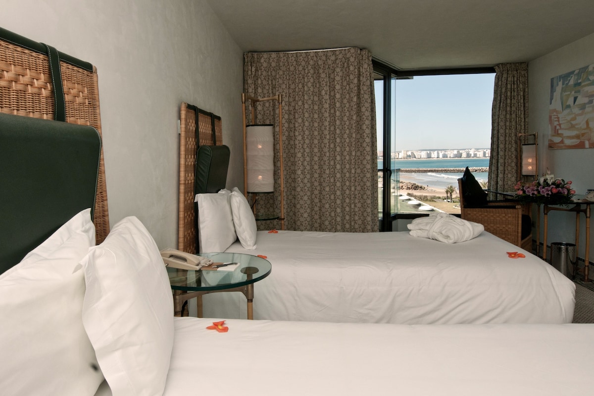 A hotel room with two beds, a glass table, a telephone, and a view of the beach and cityscape.