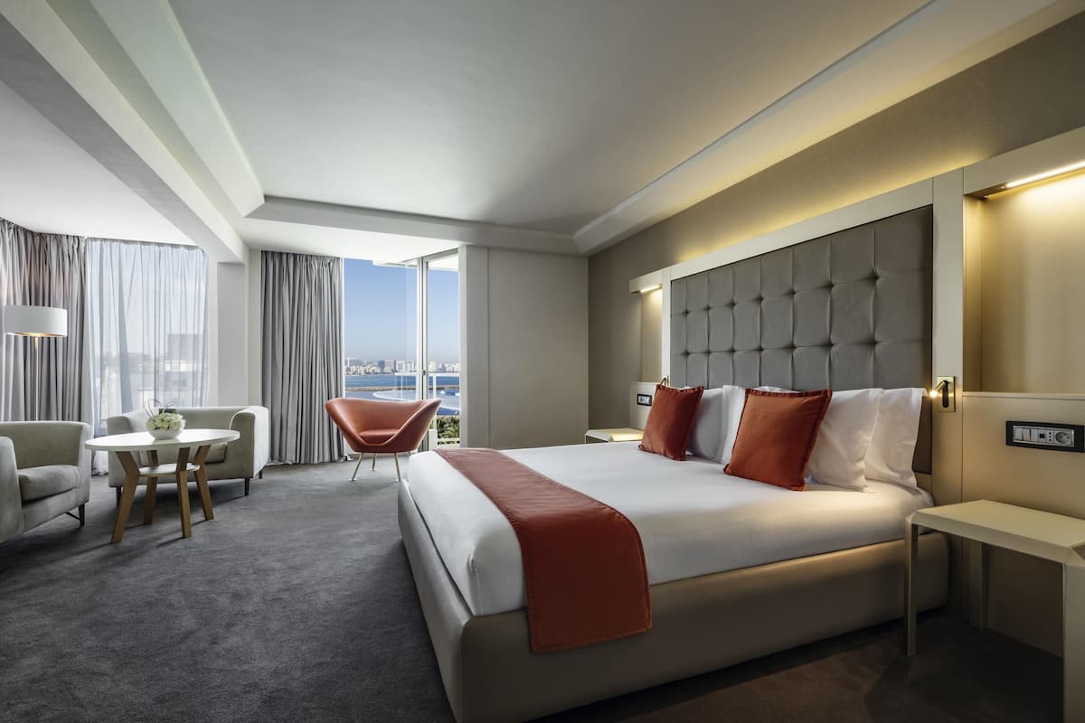 A hotel room with a large bed, a seating area with a chair and table, a bedside table, and a view of the city.