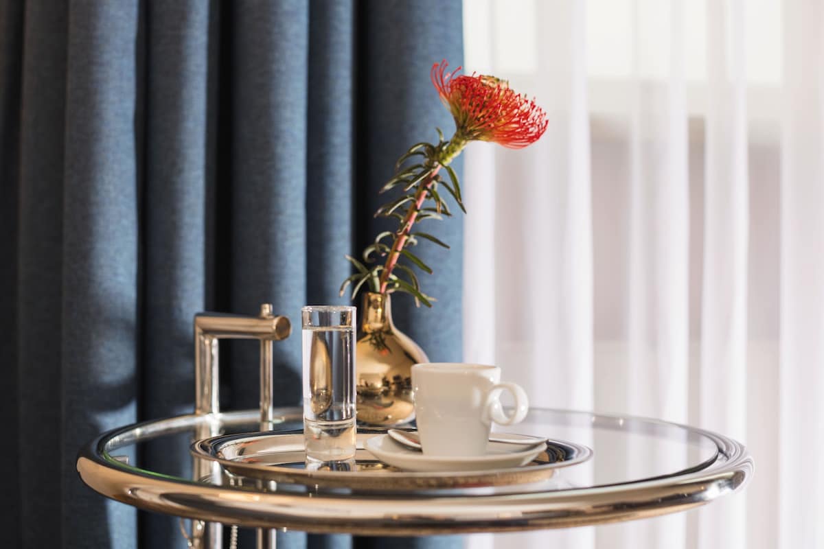 A clear glass tray with a white cup and a glass of water, a small vase with a red flower, and a gold-colored vase with a plant.