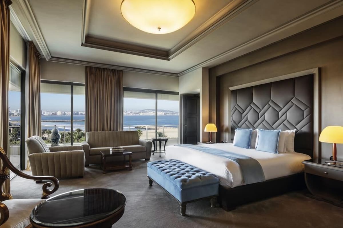 A hotel room with a large bed, a seating area with a sofa and armchairs, a small table, and a view of the city and sea.